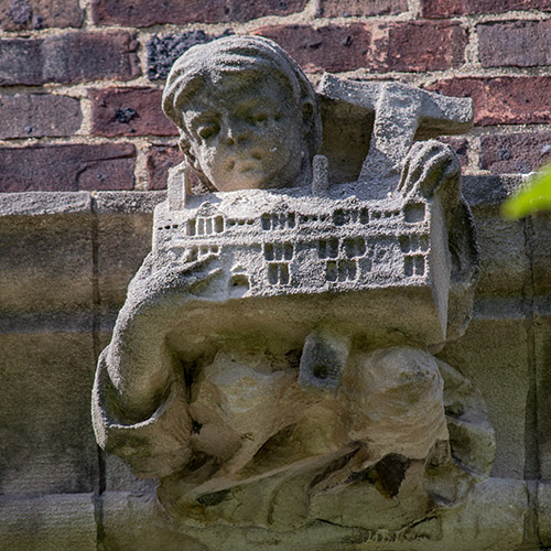 caption: In a sly reference to Cope and Stewardson’s role in designing the bosses and grotesques of the Quad, this figure holds a miniature representation of one of the Quad buildings. 