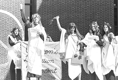 caption: Five women students in togas in front of the stainless steel Peace Symbol sculpture at Van Pelt-Dietrich Library, on March 17, 1970, during a Penn student protest promoting the Women’s Liberation Movement.